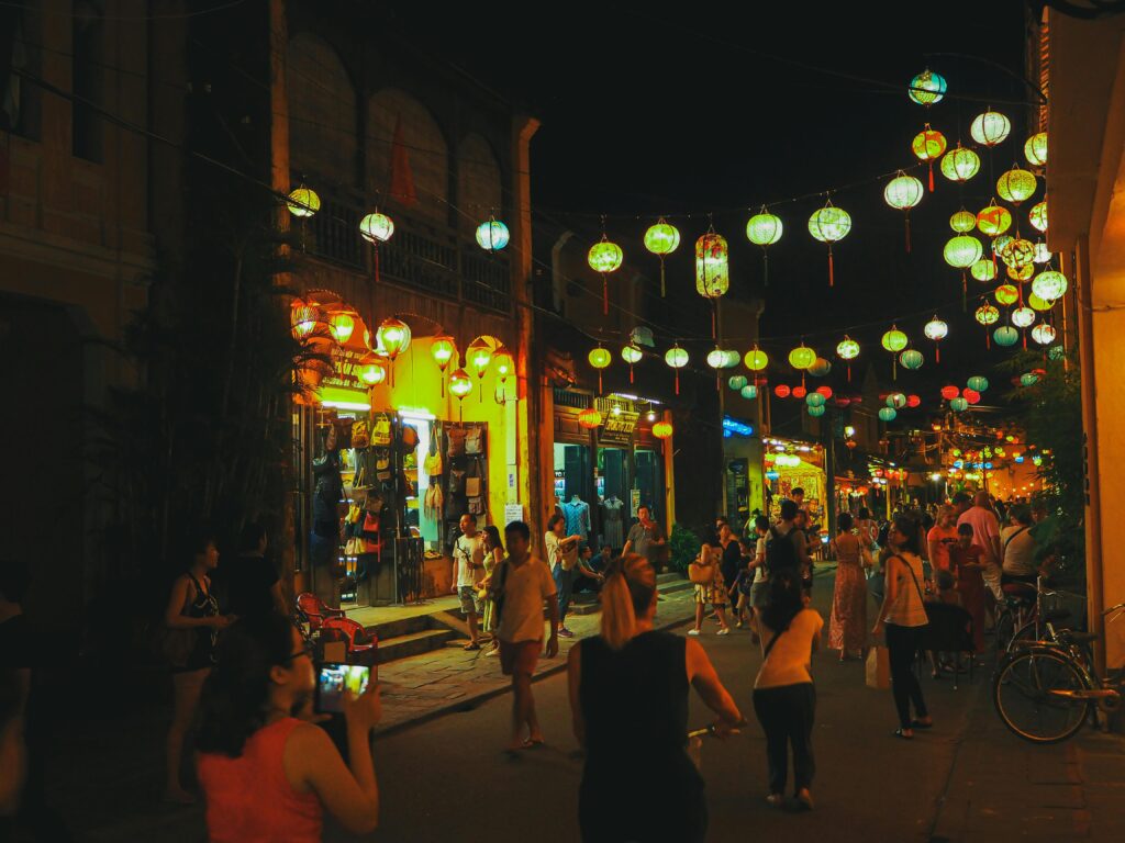 Crowd of unrecognizable diverse tourists walking on narrow pedestrian street with aged houses and cafes in city old district decorated with Hoi An paper lanterns at night